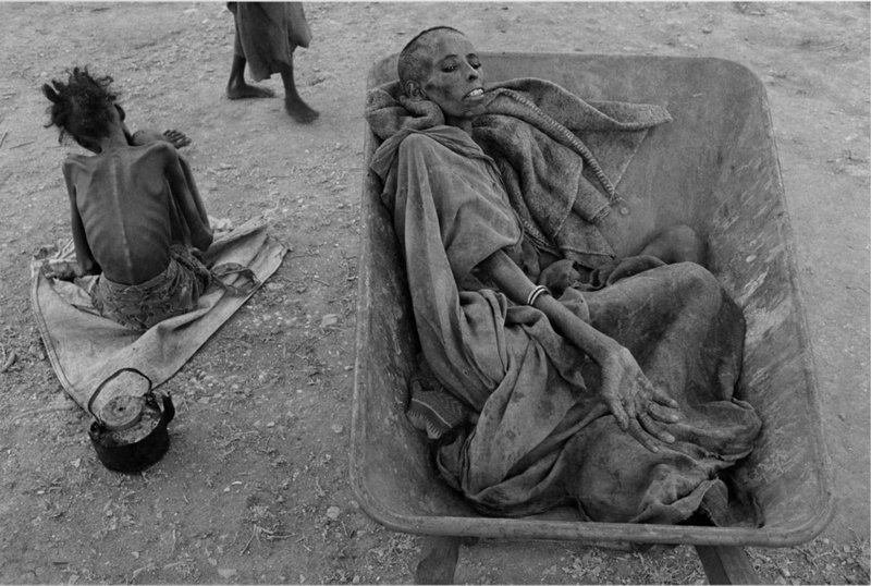 Photo taken by James Nachtwey when visiting Somalia in 1992. The photo depicts a starving woman who physically doesn’t have the strength to stand waiting to be taken to a feeding station in a wheelbarrow.