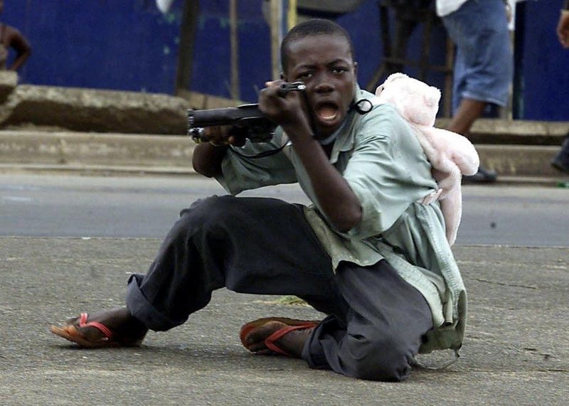 Child soldier with a teddy bear backpack aiming his rifle to the cameraman in Monrovia during the second Liberian civil war, 2003