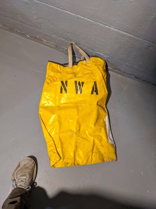 Yellow NWA Zipper Bag hidden in wall of basement. Approximately 3 feet tall with heavy black zipper on top.

Looks like a ballast bag. Filled with Sand up to 50lb. Usually needed when there are too few passengers on a flight. Those weights are used to balance the overall weight of the flight to optimize fuel usage and prevent catastrophic situations.