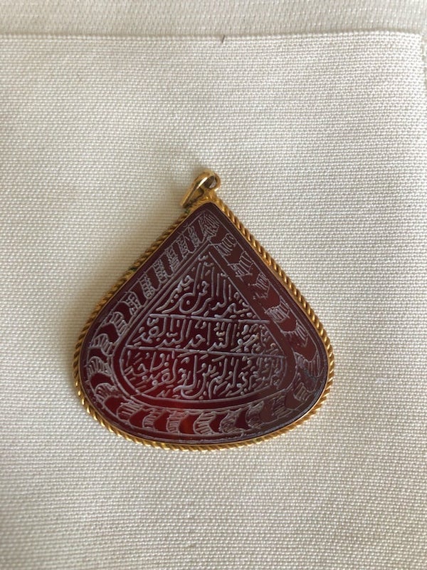 Small, pendant-like, carved into some kind of reddish material. About an inch across or two. What are these?

An Ayatul Kursi necklace. The Ayatul kursi is 255th verse of Al-Baqara Sura, the second chapter of Quran.