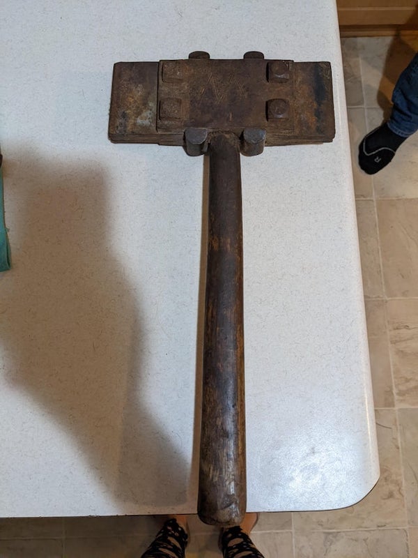 Heavy metal object made up of several plates bolted together on a wooden handle. Some markings on either side including what we think is the letters ‘C’ and a large ‘W’.

A: It’s a type of bush hammer used in stone work.