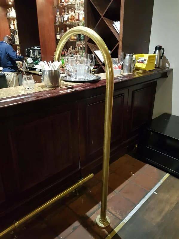"I’ve seen this cane-shaped (usually) brass post at numerous bars and pubs and wondered what it is and what it is used for. l have been unable to Google the correct sequence of words to get a result."
<br/><br/>
"A “divider” or “service divider”. It serves to keep standing customers to one side, away from the part of the bar where servers pick up drinks. Also notice the opening under the bar, the bartender (in some designs) can lift the top and gets in and out. You don’t want customers standing there blocking access."