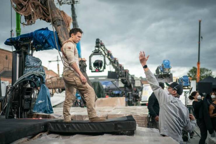 behind the scenes movies - Tom Holland stands on a big, comfy-looking mat, listening to director Ruben Fleischer on the set of Uncharted (2022).