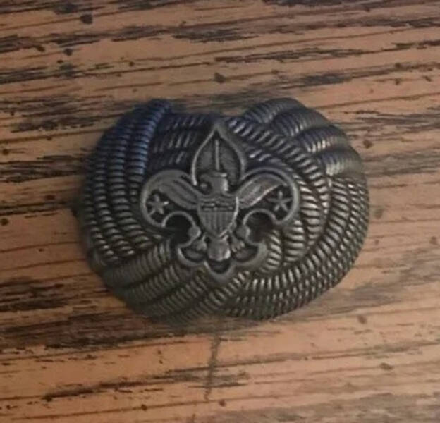 “A small metal trinket found buried on a rural farm — it appears to have an eagle, Flor de Lis, and rope pattern.”

"It’s a Boy Scouts neckerchief slide."