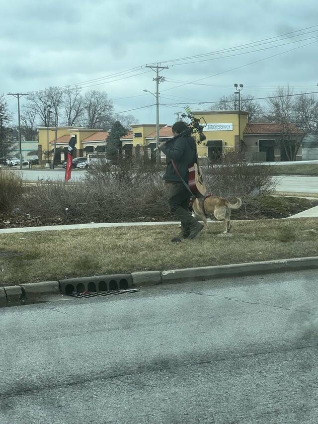 "Guy walking around with crossbow, guitar, and dog"