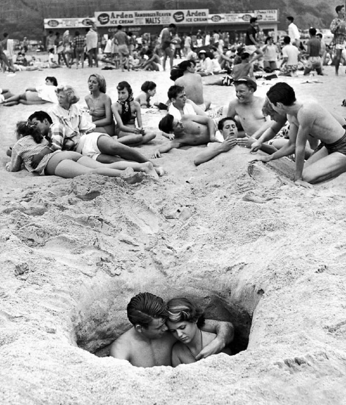 Couple Cuddling While Sitting In A Hole As Others Enjoy The Beach On The 4th Of July In Santa Monica, California, 1950. (Photographed By Ralph Crane)