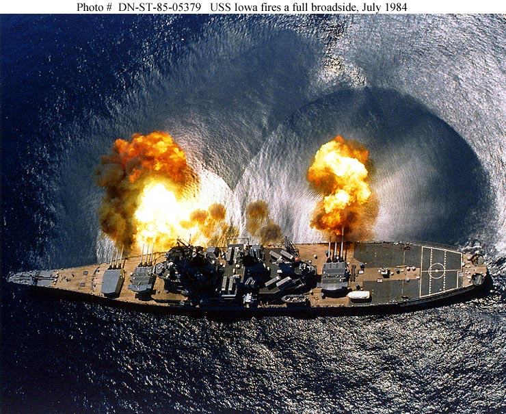 An aerial view of a battleship firing its main guns.