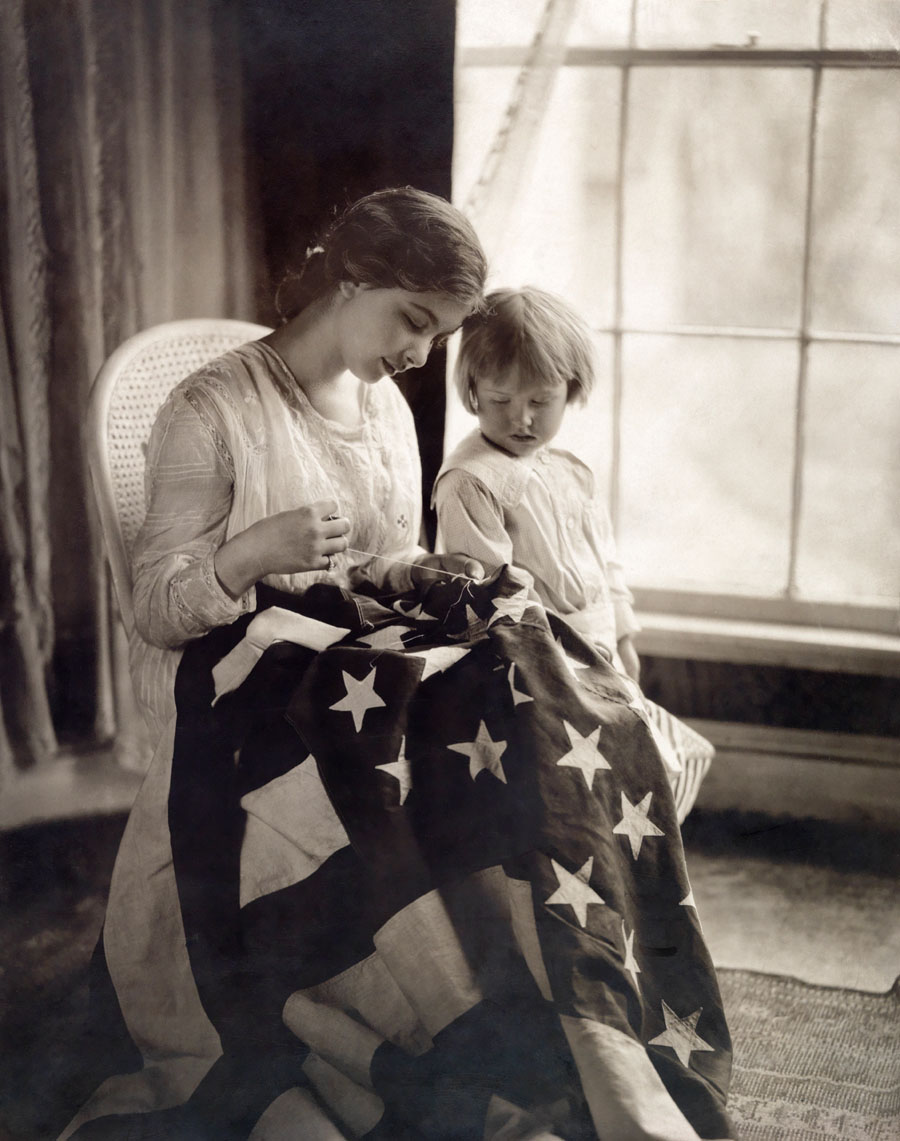 A child looks on as stars are being carefully sewn to a US flag, 1917.