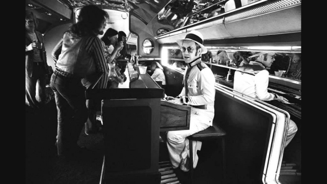 Elton John playing the piano in the bar of his private jet, 1976.