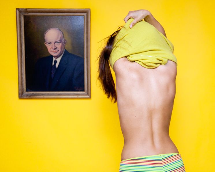 Picture of woman taking off her shirt in front of a portrait of a man.
