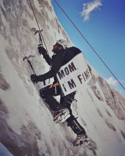 Funny pic of a mountain climber using ice picks to climb a very steep slope, holding a sign saying MOM I'M FINE.