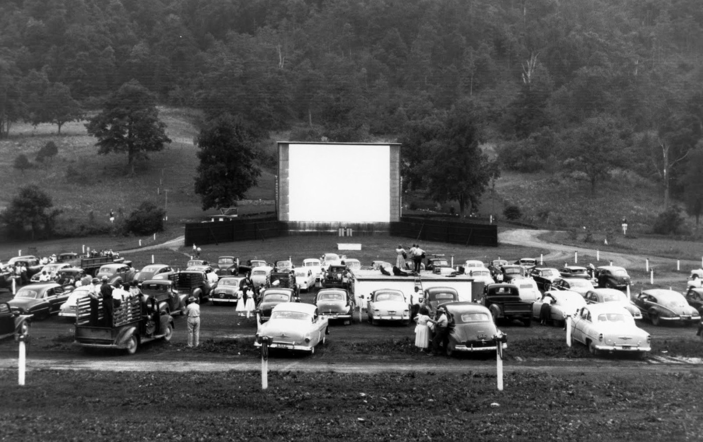 A drive-in movie theater preparing to start its feature in Georgia, US in 1950. Each pole near the cars has a speaker people can use to hear the film, and in this theater there is a band on top of the reel building doing live music.