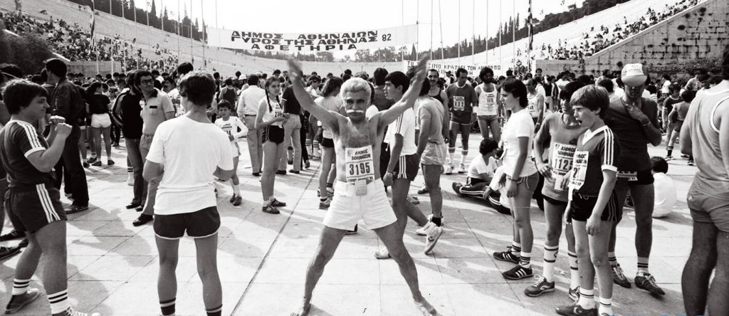 An older man playfully poses for the camera prior to some organized public races in Athens, Greece in 1982. The young man on the right seems caught quite off guard.