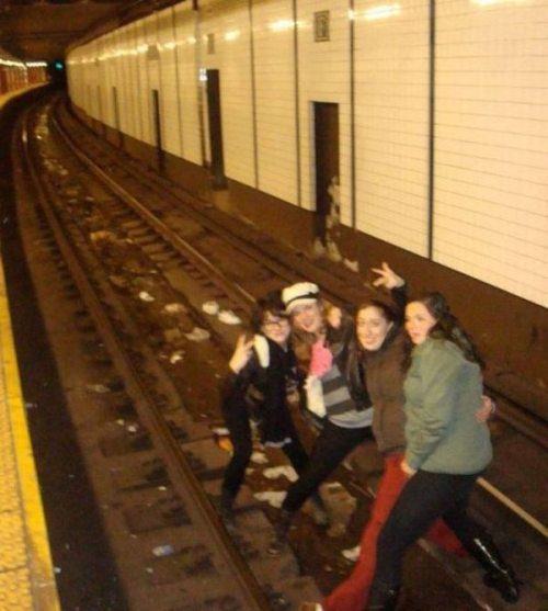 Girls posing for a pic on the train tracks of the subway, with oncoming train visible.