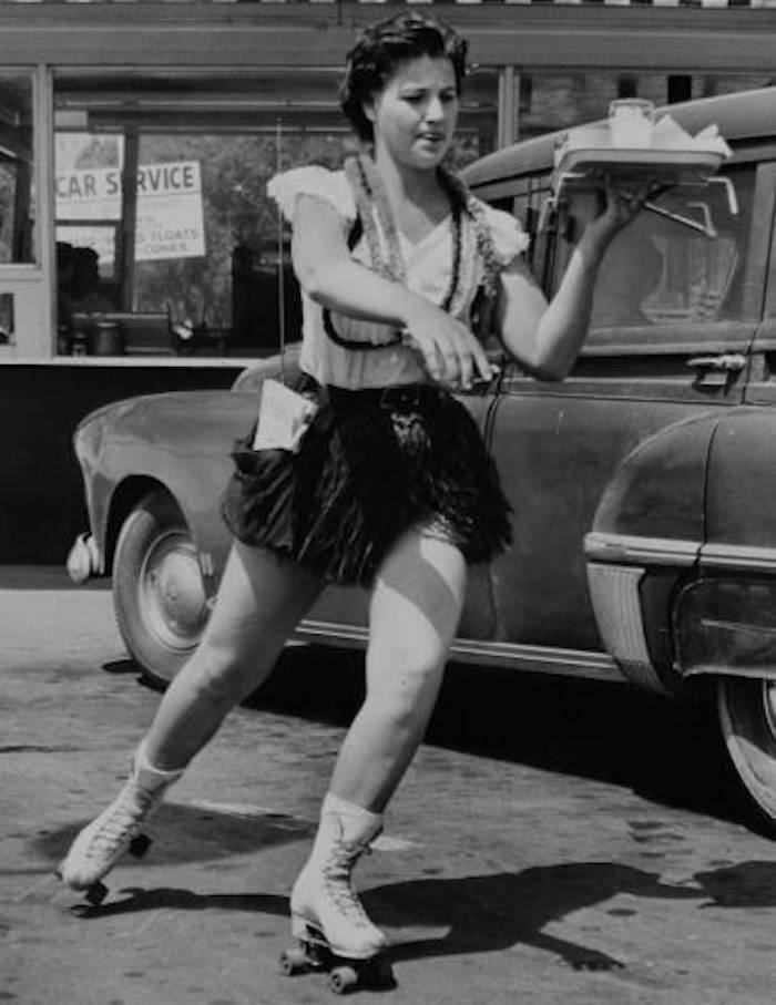 A young lady works as a waitress for a drive in restaurant somewhere in the US in the 1950s. These servers with this style of waiting on people, which was by bringing the patrons foods directly to their cars, were known as carhops. Sometimes, as pictured, they wore roller skates to move quicker. It started in the 1940s and some novelty restaurants still have them today, but they most died out in the 1980s. They were really big in the 1950s and 1960s, and the outfits went from full body uniforms in the early days to short shorts and mini skirts by the 1950s, adding sexual intrigue to a rather common job.