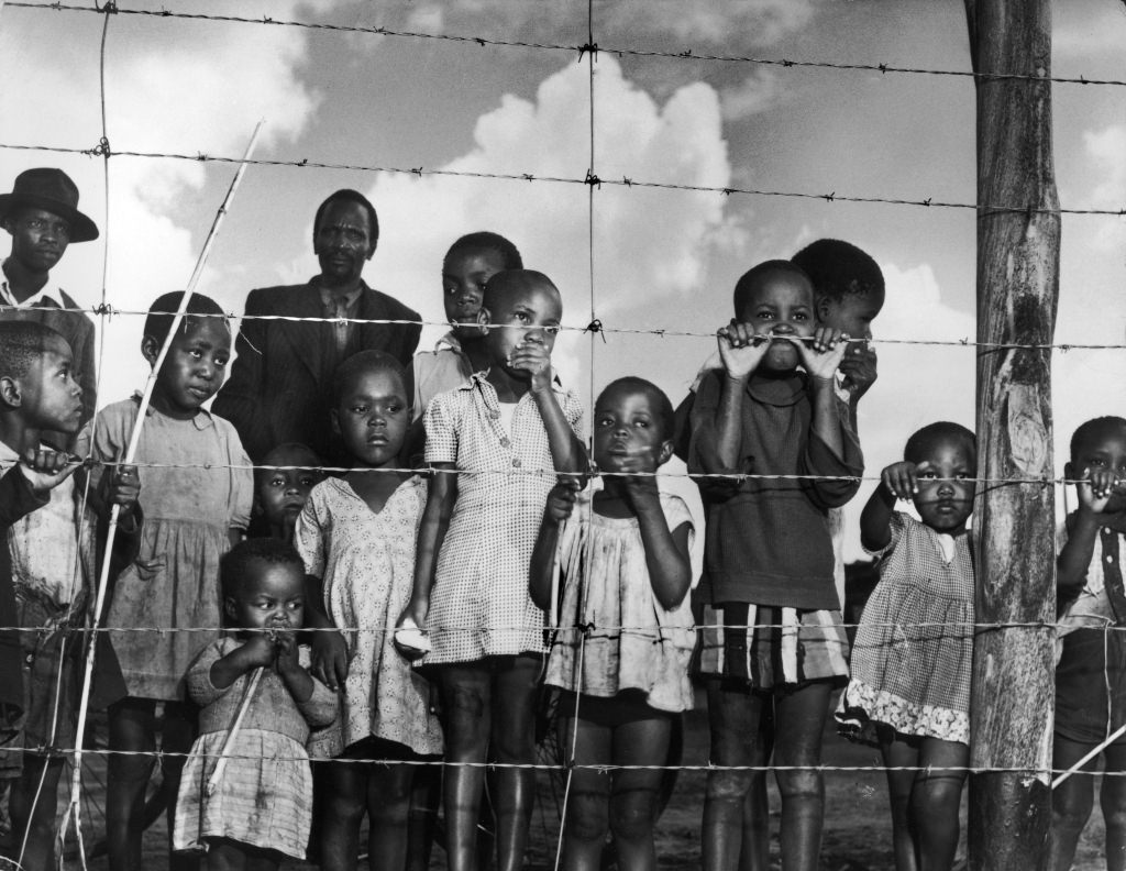 Mostly children stand near a fence that marks the end of a shantytown in South Africa in 1950.