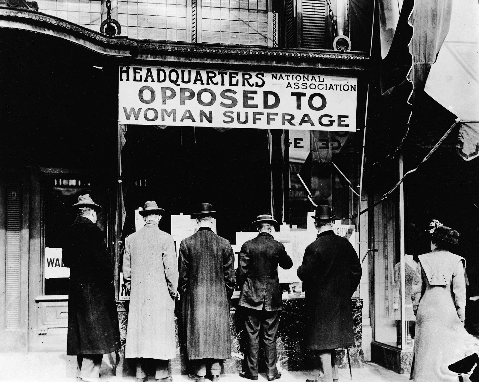 A group of men read literature posted in the window of the National Association Opposed to Woman Suffrage in New York City, while a woman stands off to the side, 1910.