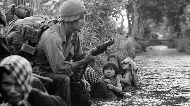 Women and children hiding in a stream look on as American Marines move past them in Vietnam, 1967.