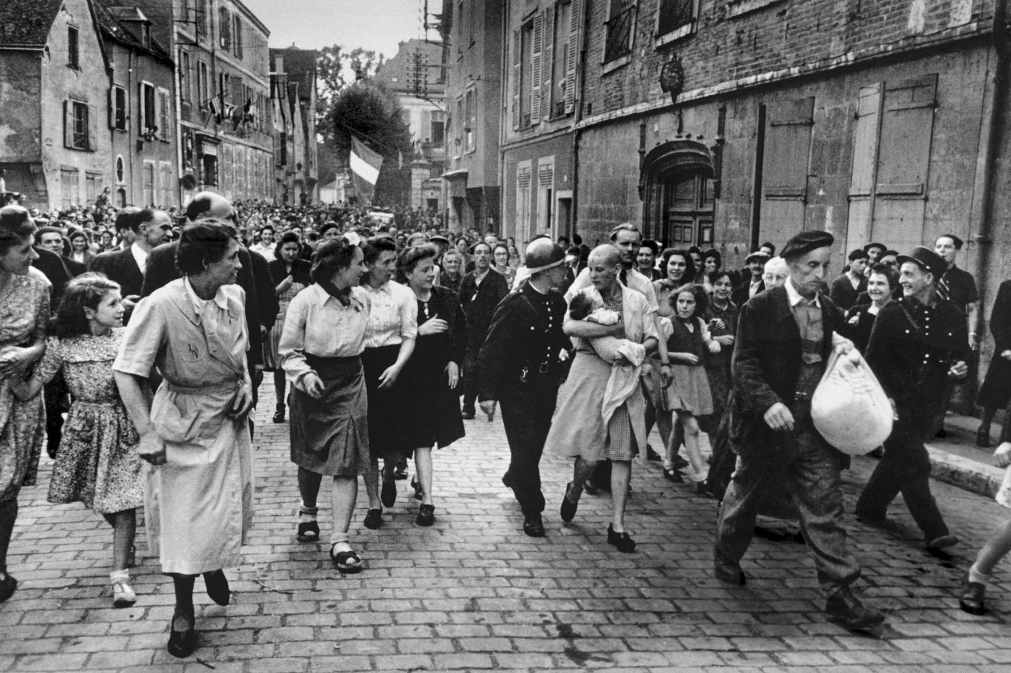 A women with her baby fathered by a German is escorted down the street and out of town by an angry mob after she had her hair cut off and was humiliated in a town in France, 1944.