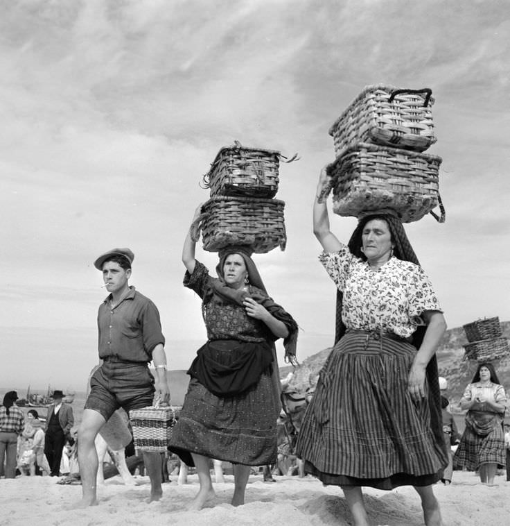 People carrying things at the beach in Portugal in 1954. Notice the woman in the background on the right not even holding the baskets on her head up with her hands.