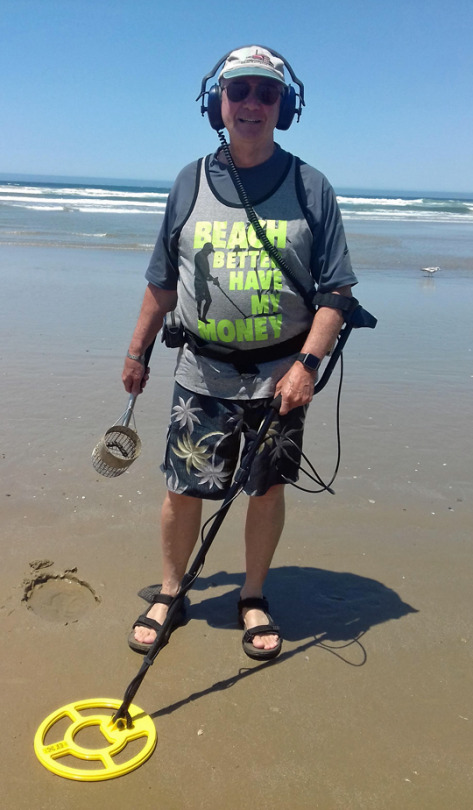 Man at the sea side with a metal detector and shirt that reads 'BEACH BETTER HAVE MY MONEY'
