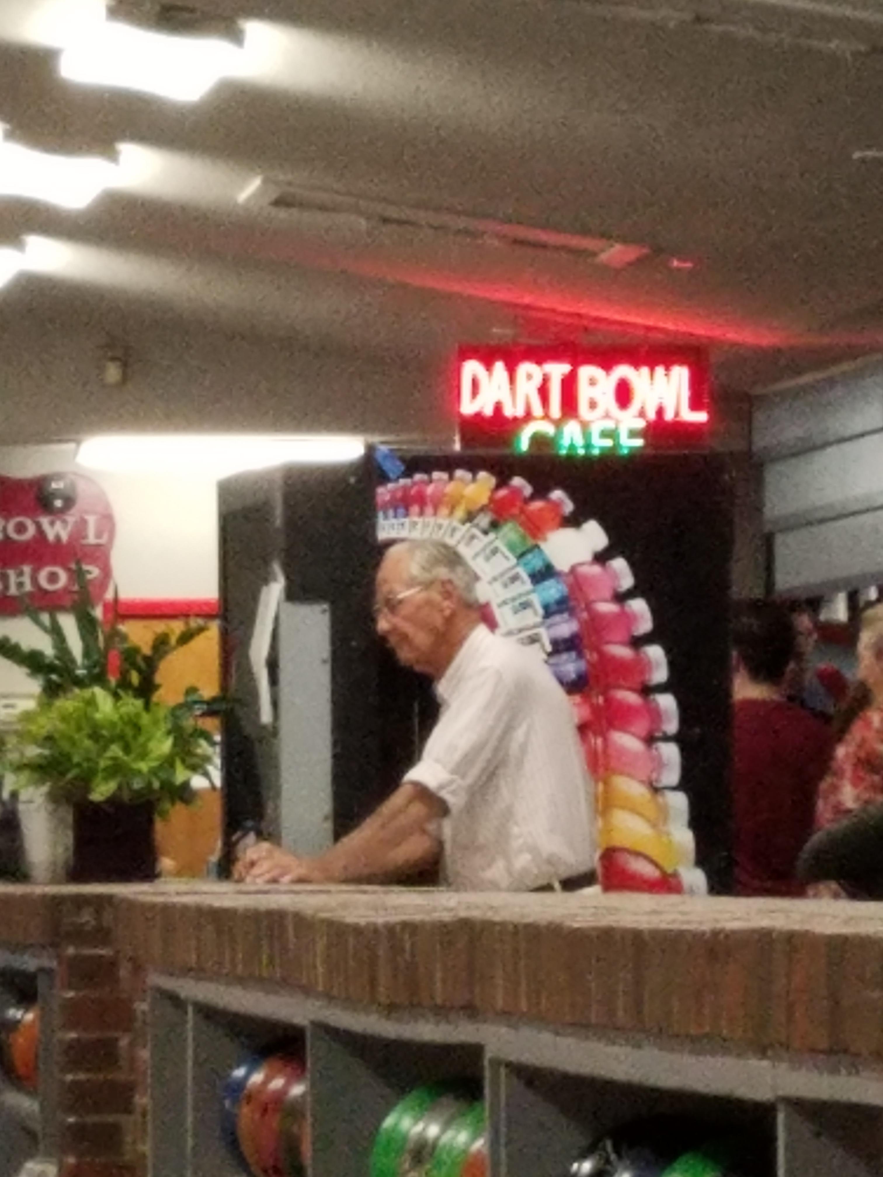 Old man at the counter in front of a vending machine that makes it look like he is wearing an awesome Indian head-dress of Gatorade instead of feathers.