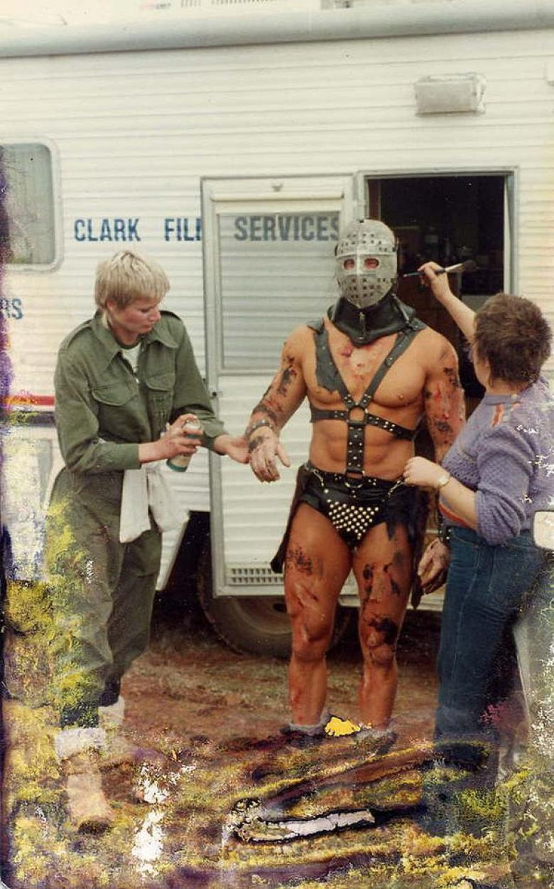 Picture from behind the scenes of Road Warrior of muscled character getting make up put on by some trailer