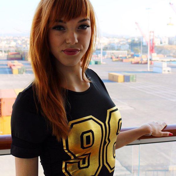 Pornstar Anny Aurora wearing a black shirt with gold 98 printed on it, standing on a balcony looking out over a shipping yard