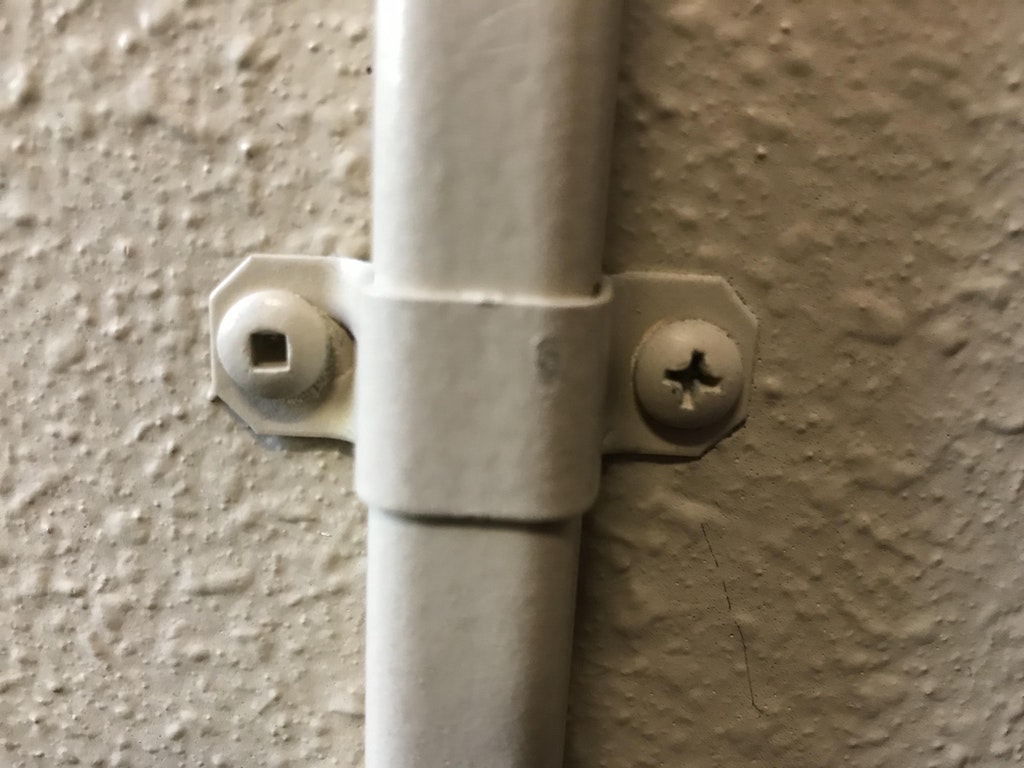 infuriating clamp securing a pipe conduit to the wall using two screws of differing variations, with one being a square key design and the other the Phillips plus configuration