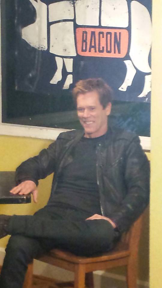 Kevin Bacon sitting under a sign that says Bacon