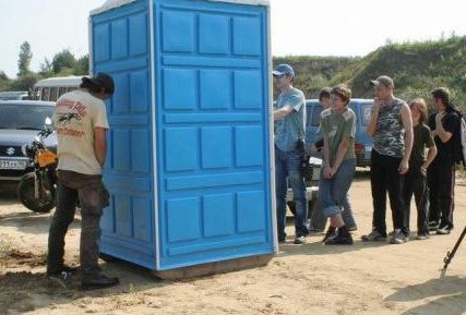 Picture of a line of people waiting at a porta-potty and one guy takes matters into his own hands.
