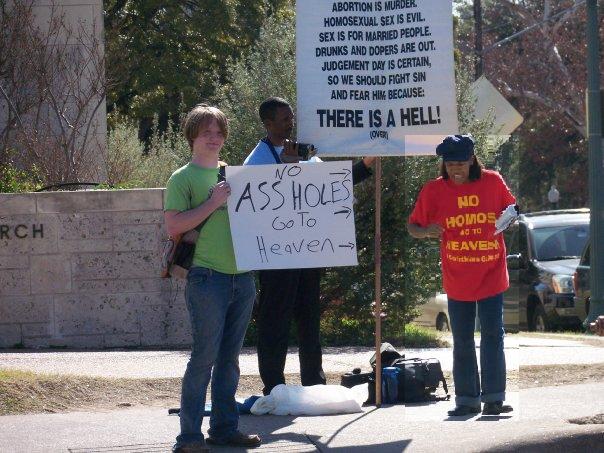 A woman who has a shirt that says No Homos Go to Heaven has a kid protesting her. His sign says No Assholes Go to Hell with arrows pointing at her.