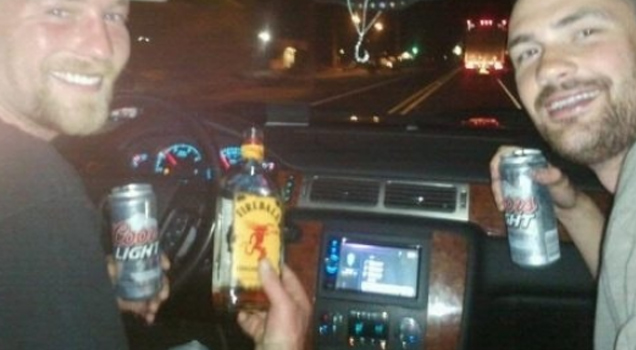 two guys--one in the driver's seat of a car; one in the passenger's seat--hold 3 bottles of alcohol and smile at the camera, which is in the opposite direction of where the car is headed. There is a bus 30 yards ahead of them.