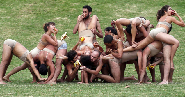 People on grass in flesh-colored outfits writhe on each other while eating apples.