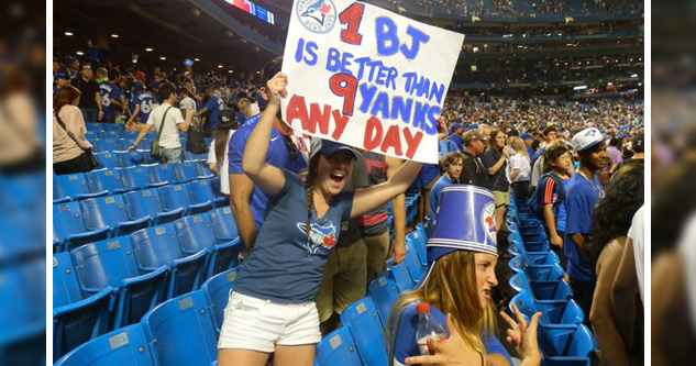 Woman in Blue Jays clothes holds a sign saying: 1 BJ is better than 9 yanks any day.