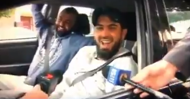 Two men smiling in a car talk into microphones held by interviewers.