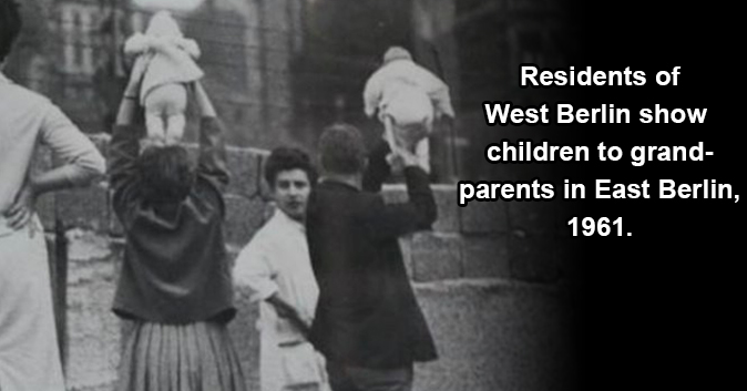 Black and white photo of two people holding two babies over a wall. Text: Residents of West Berlin show children to grandparents in East Berlin, 1961.