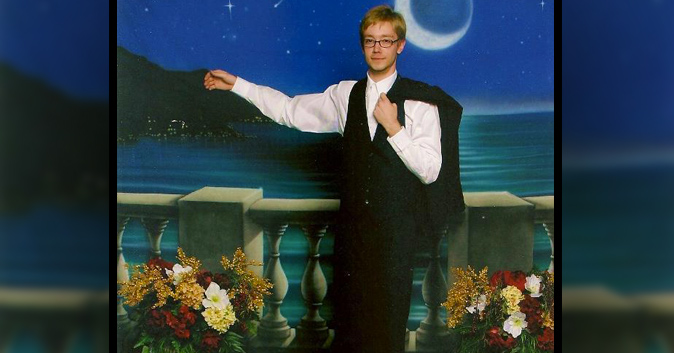 Guy in a suit stand for a prom photo alone. His arm is around a pretend woman.