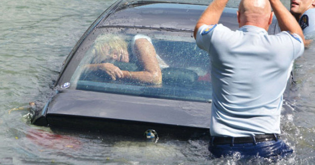 A woman gasps for breath in the back of a car that is sinking into the water. Two police officers struggle to get into the car to save her.