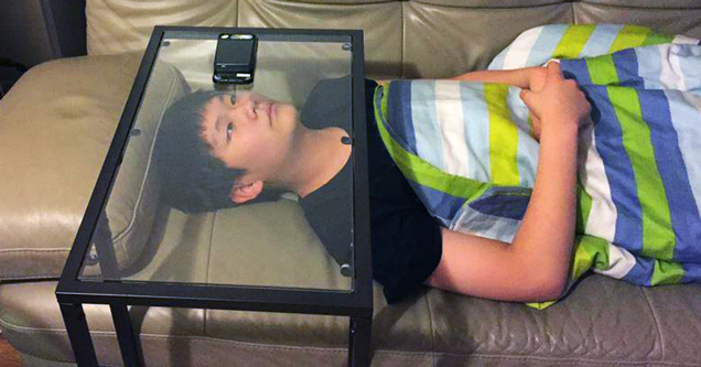 kid watching his phone while laying under a glass table