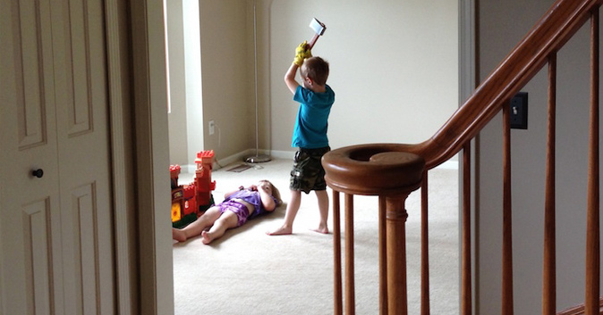 Kid holds a toy axe over his sister, who is laying on the carpet.