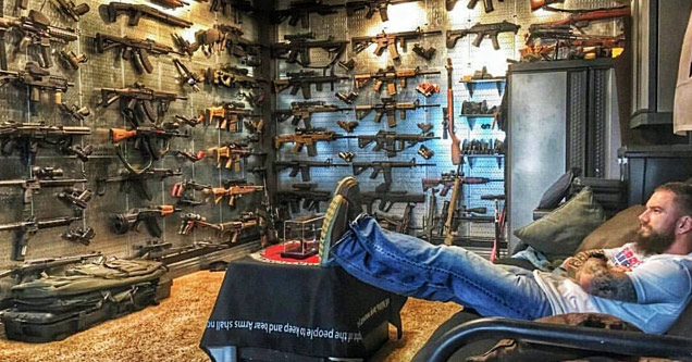 man sitting with legs on table in a room full of guns