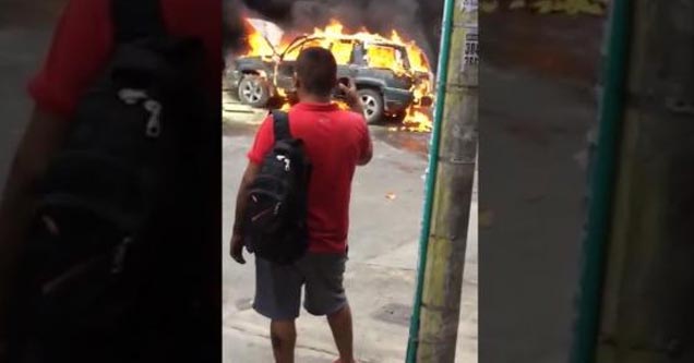 a man in a red shirt watching a car burn