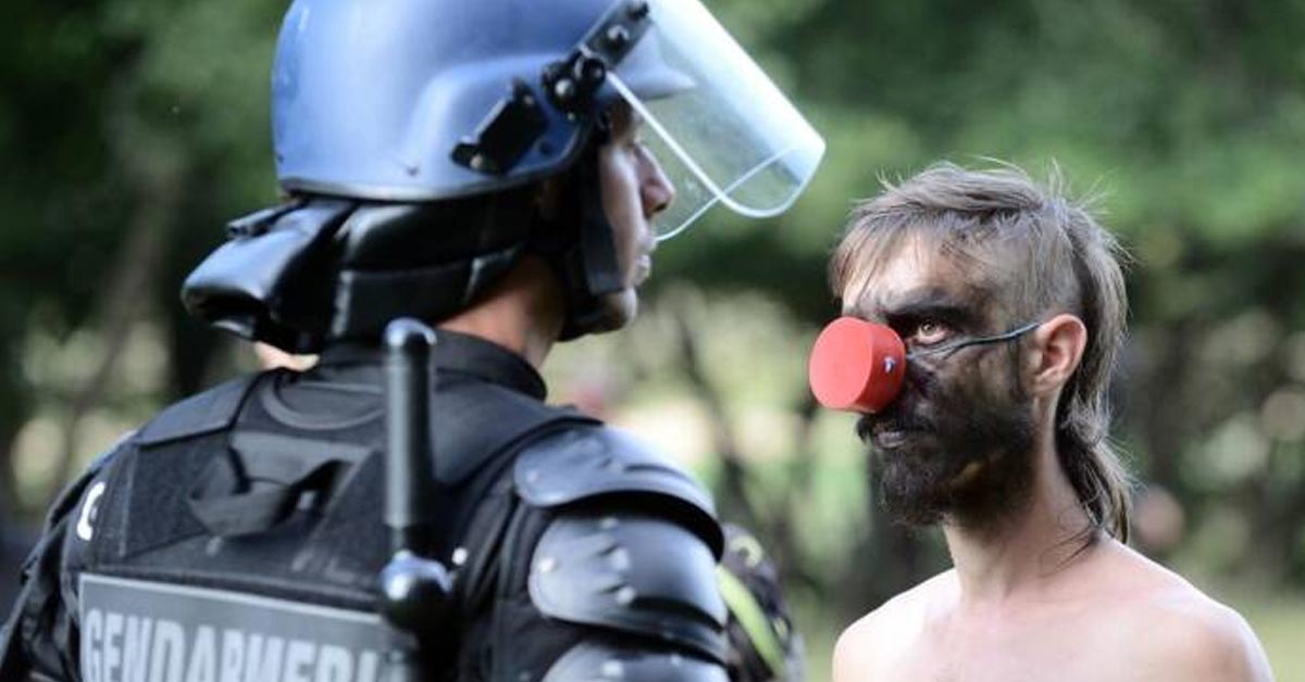 a guy dressed as a clown looking at a cop in riot gear